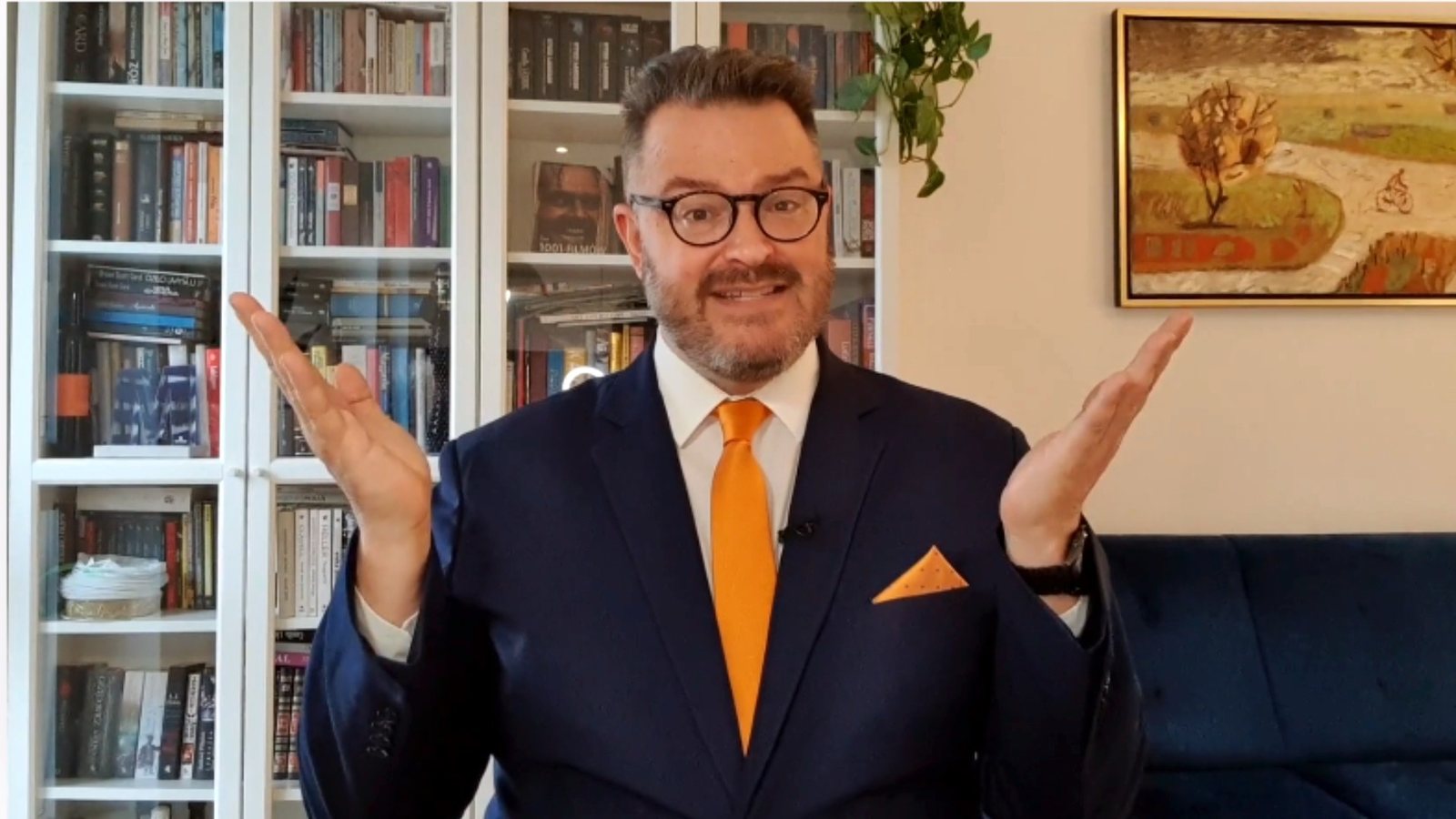 Derek Zardus teaching in front of his bookshelves, in a navy suit and orange tie